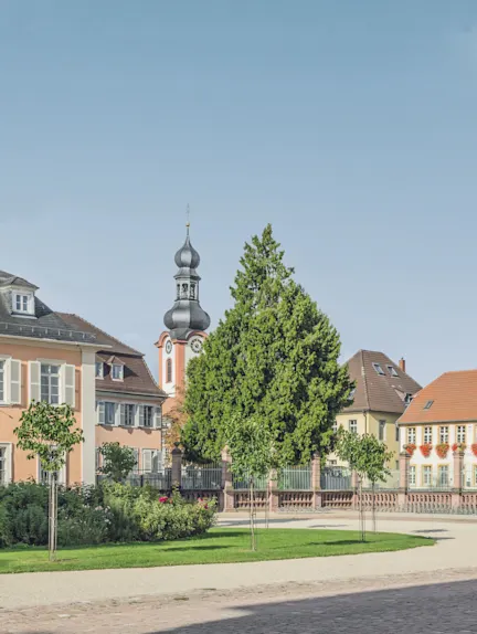 Mehrere Wohnhäuser und ein Kirchturm hinter einem begrünten, gepflegten Vorgarten.