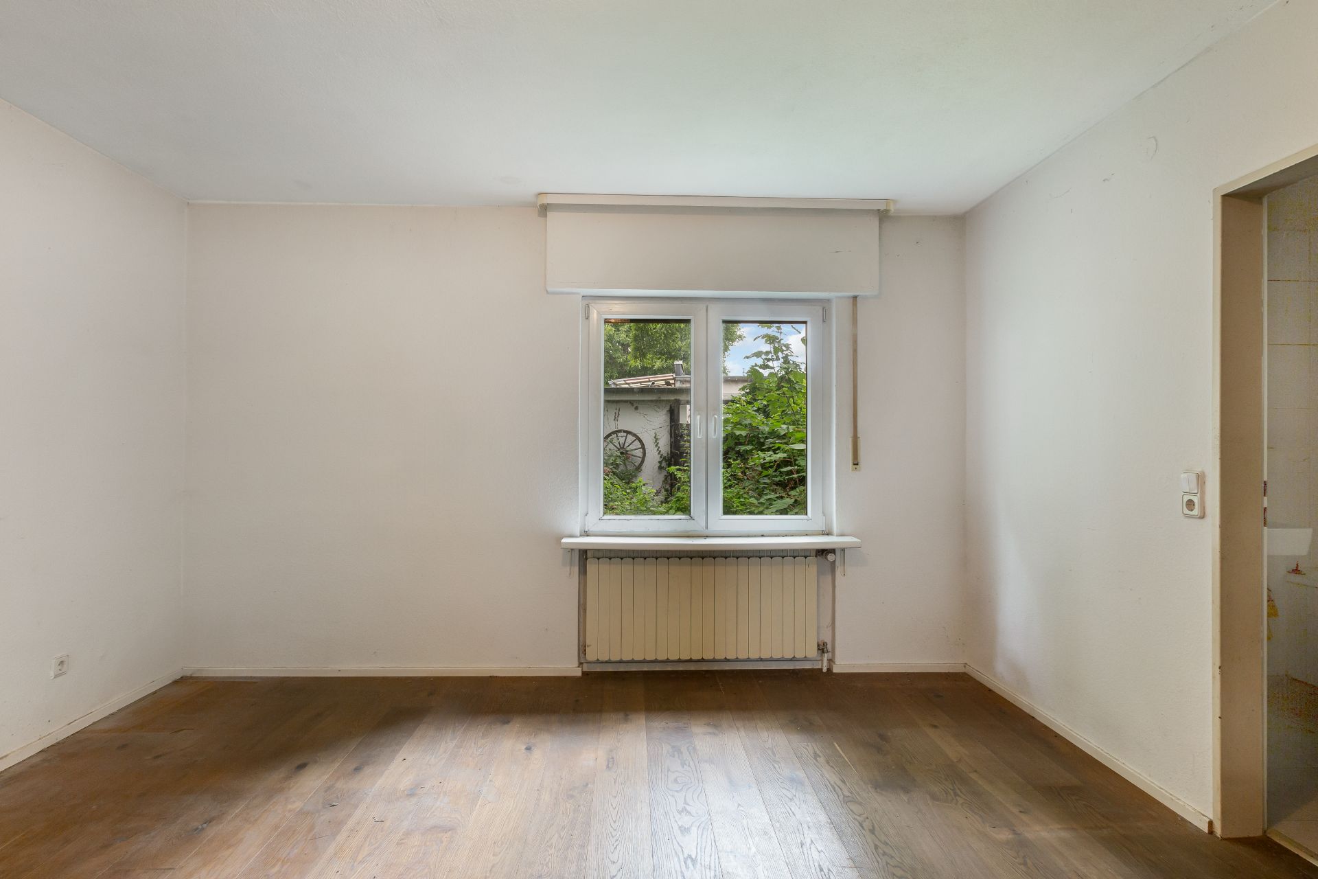Leeres Zimmer mit Holzboden, weißer Wand und Fenster mit Blick ins Grüne.