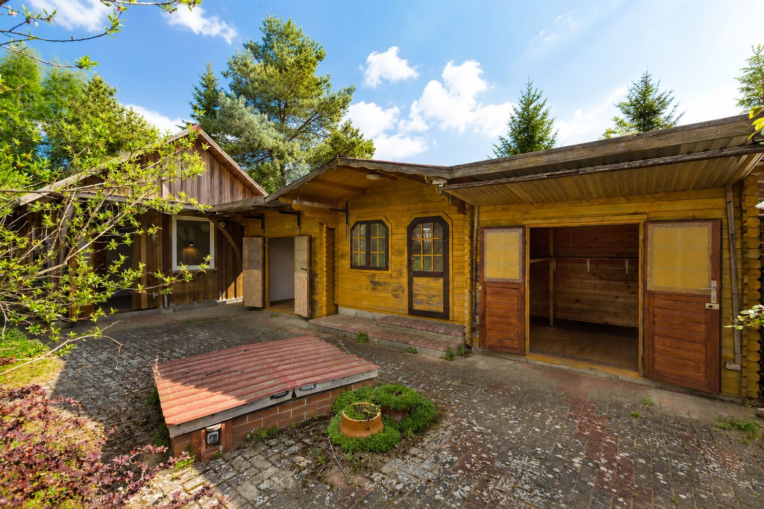 Holzhaus mit offenem Tor, Ziegelpflaster und Bäumen im Hintergrund.