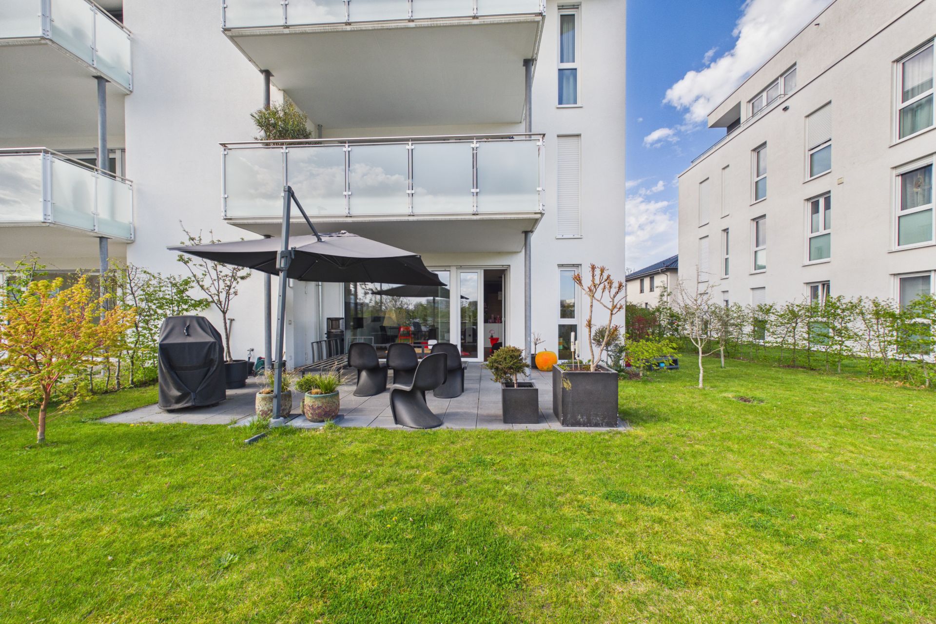 Moderne Terrasse mit Sitzgruppe, Sonnenschirm und Grill im Garten eines Wohngebäudes.