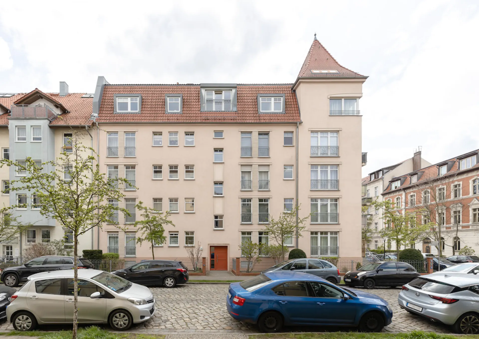 Mehrstöckiges Wohnhaus mit rotem Dach, Bäumen und geparkten Autos davor.