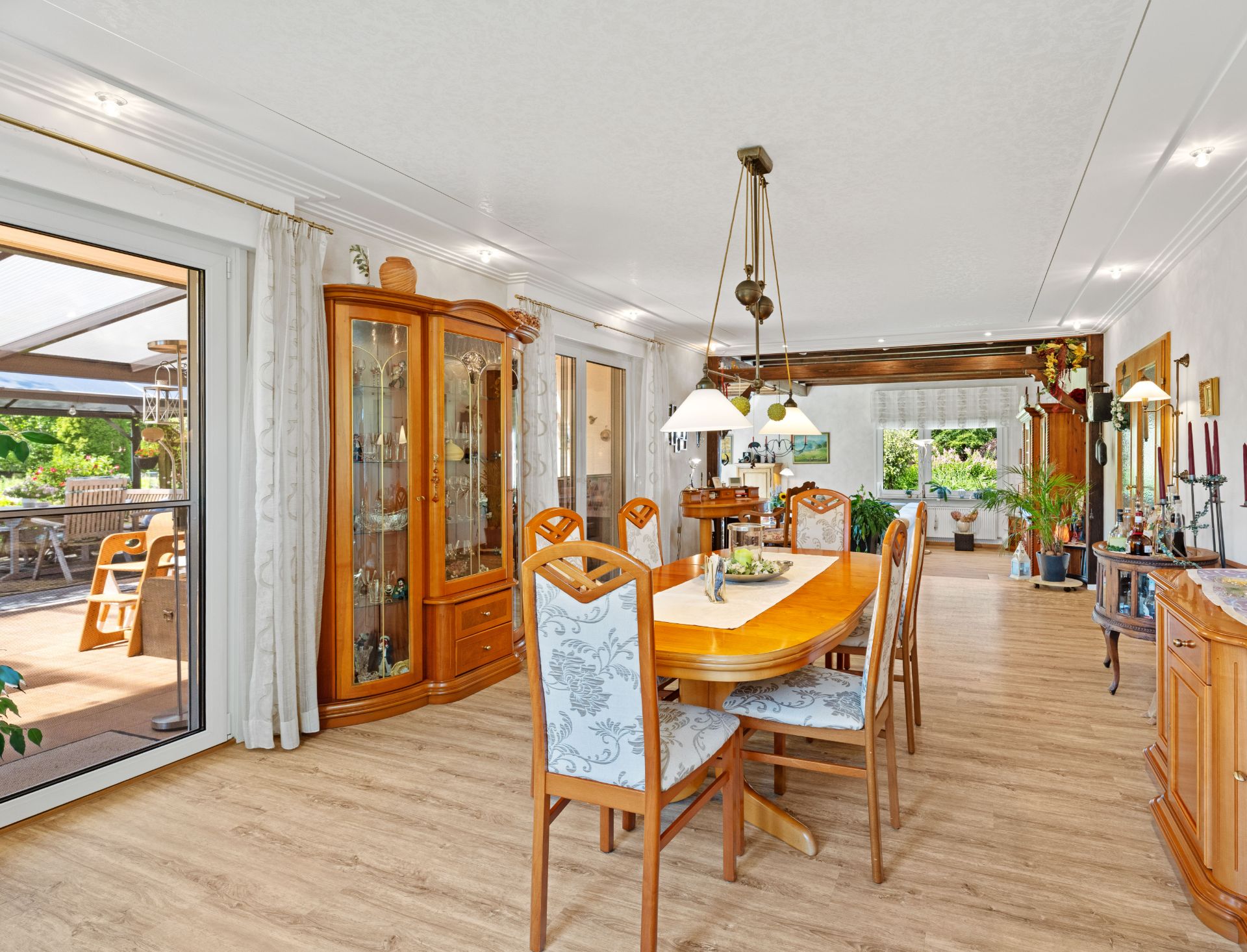 Helles Esszimmer mit Holzmöbeln und großem Esstisch, Zugang zur Terrasse, Holzboden und Vitrinenschrank.