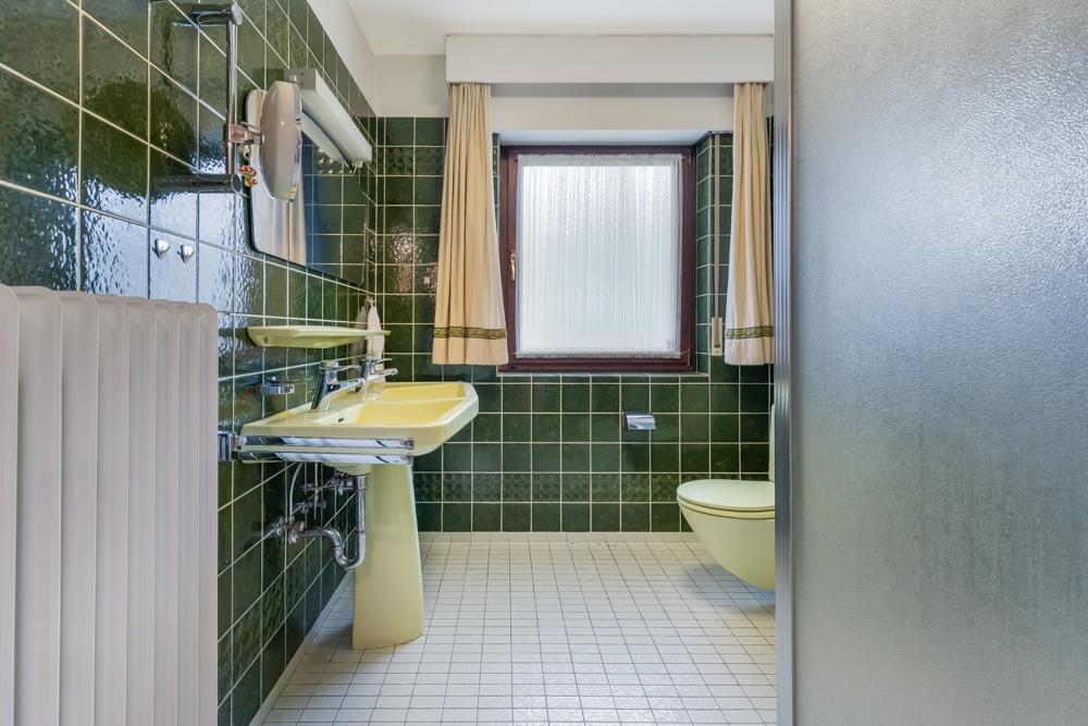 Badezimmer mit grünen Fliesen, gelbem Waschbecken, Toilette und einem Fenster mit Vorhängen.