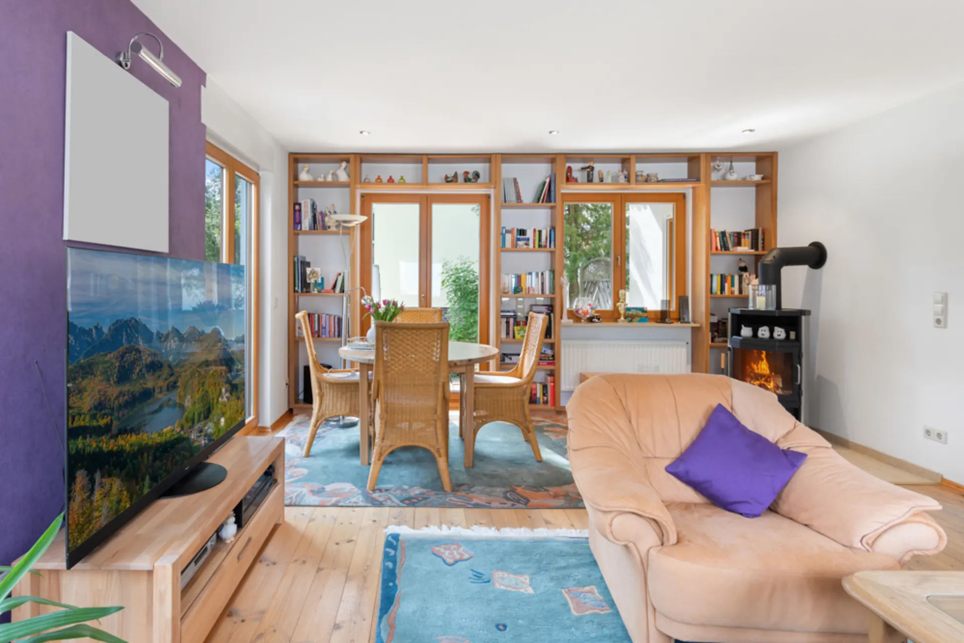 Wohnzimmer mit beigem Sofa, Kamin, großem Fernseher, Esstisch und Bücherregalen vor Fenstern.
