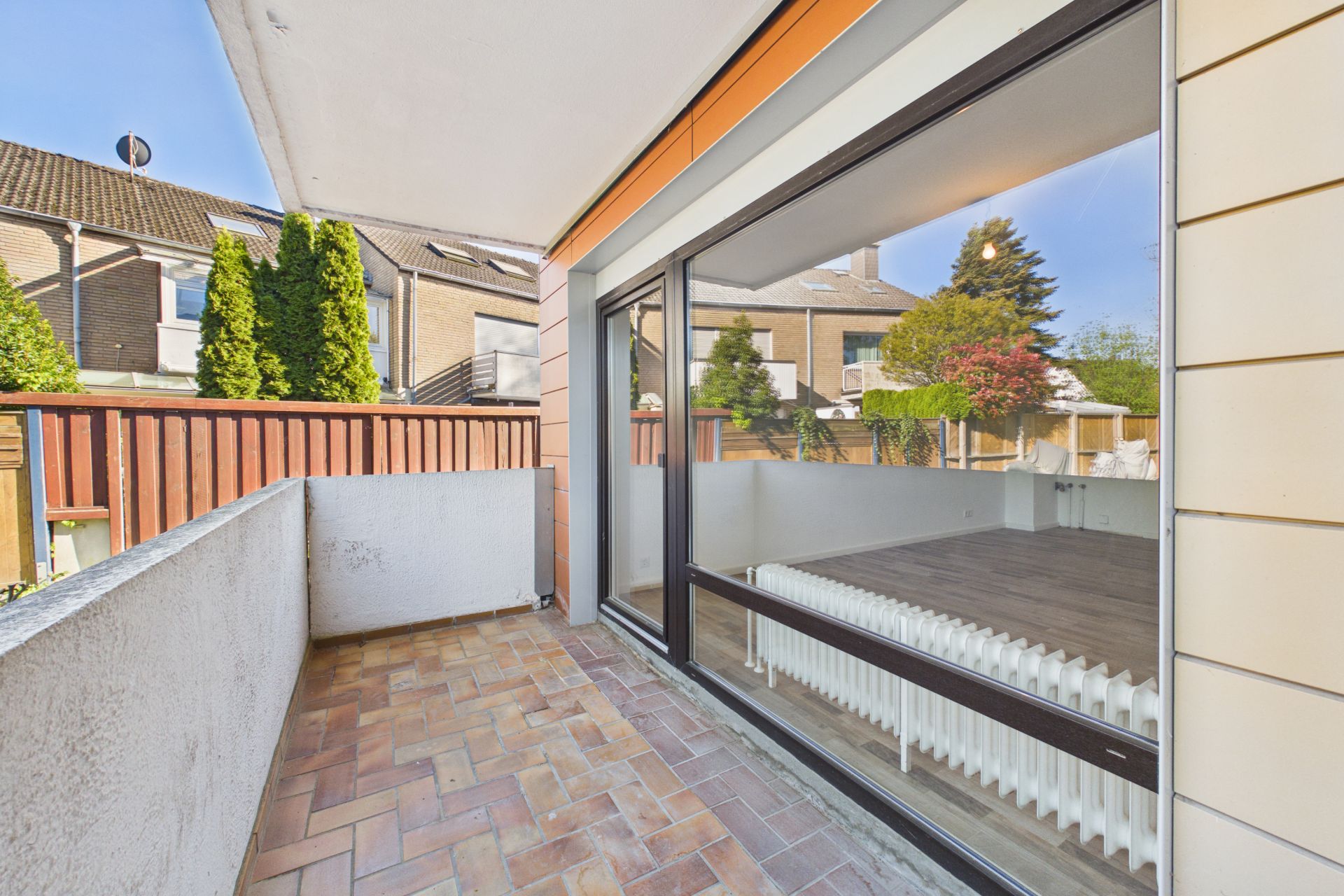 Balkon mit Fliesenboden und Glasfront, Blick in einen Garten und auf benachbarte Häuser.