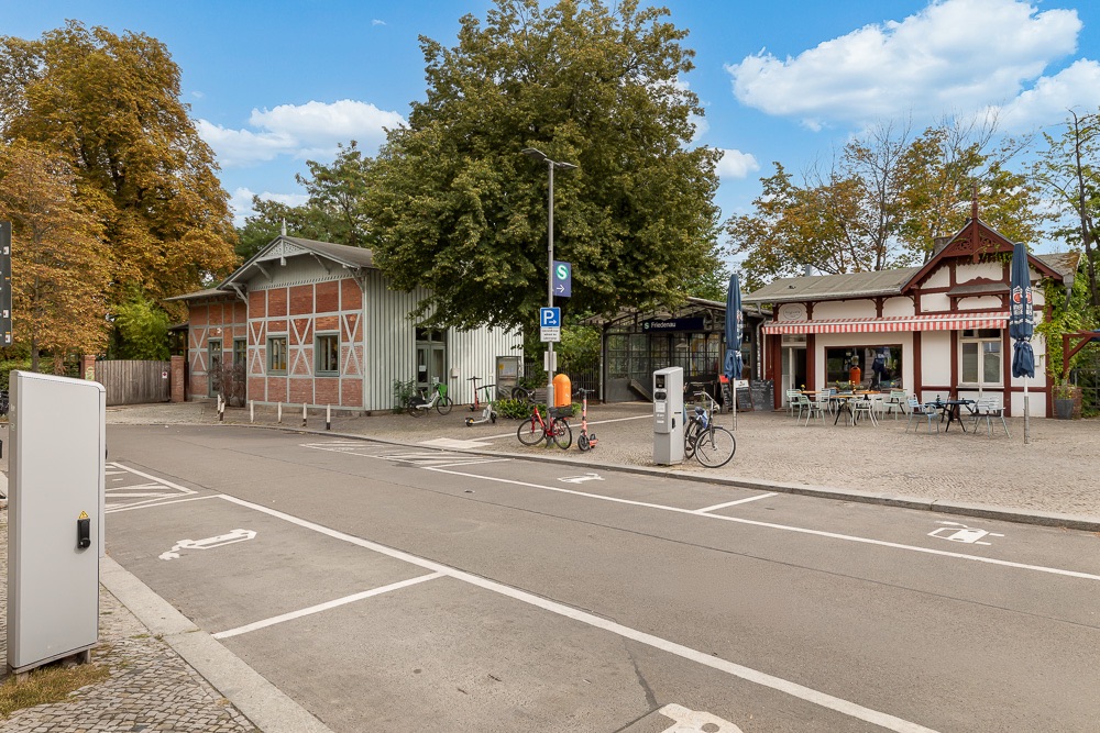 Straße mit Fahrradständern, zwei Gebäuden und Sitzbereich im Freien, Bäume im Hintergrund.