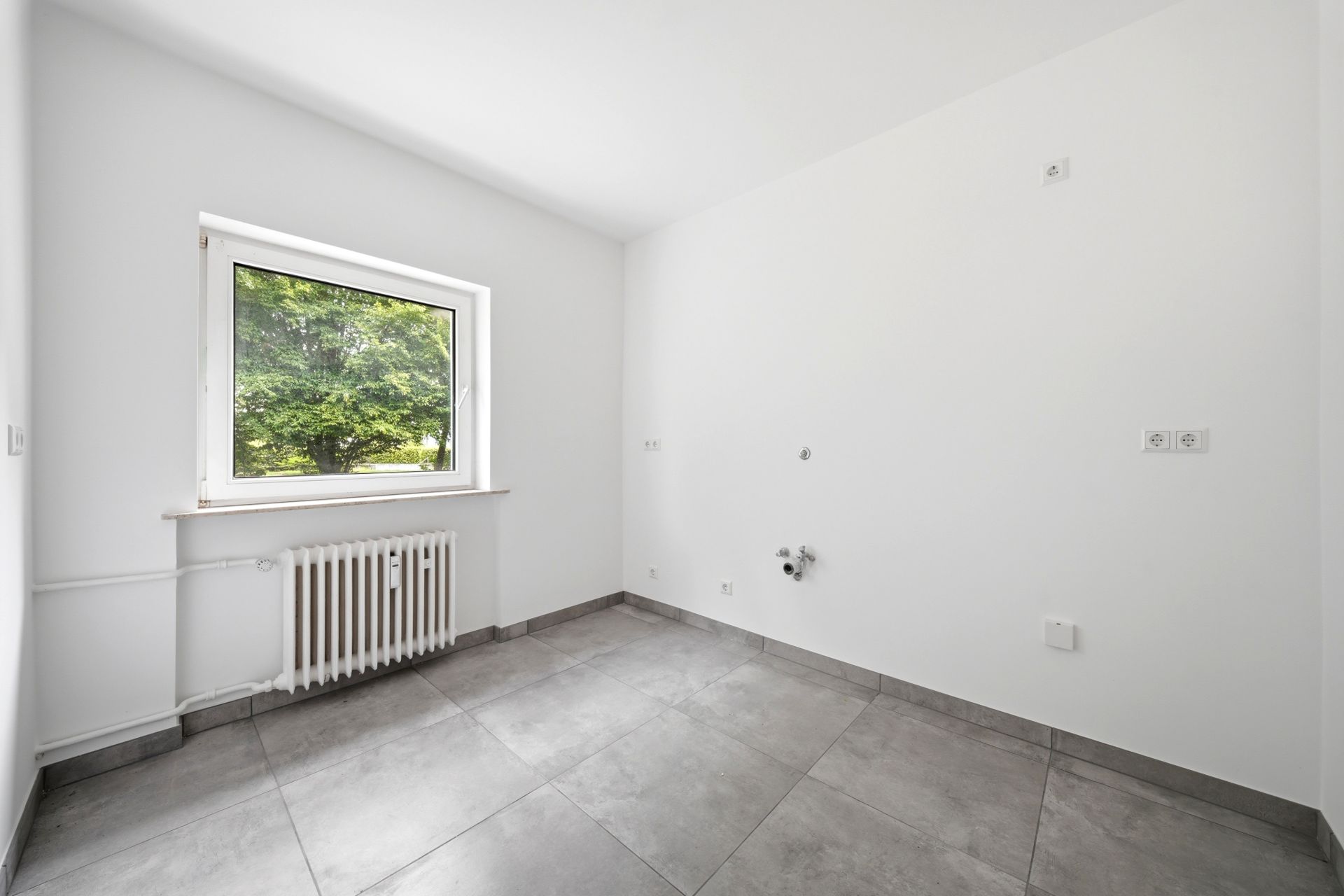 Leerer, weiß gestrichener Raum mit großem Fenster, Heizkörper und grauen Fliesen.