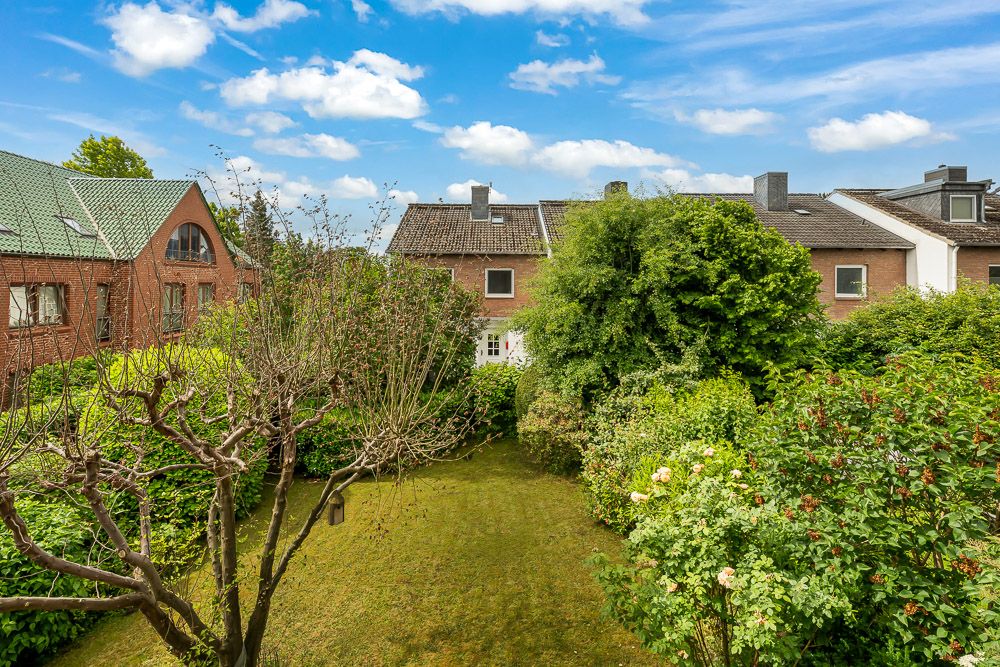 Garten mit Bäumen, Büschen und Rasenfläche, gemauerte Wohnhäuser im Hintergrund.