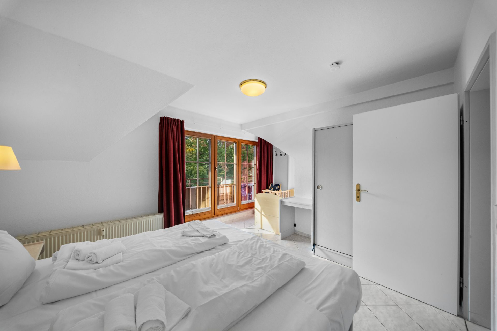 Helles Schlafzimmer mit Doppelbett, Schreibtisch, rotem Vorhang und Zugang zu einem Balkon.