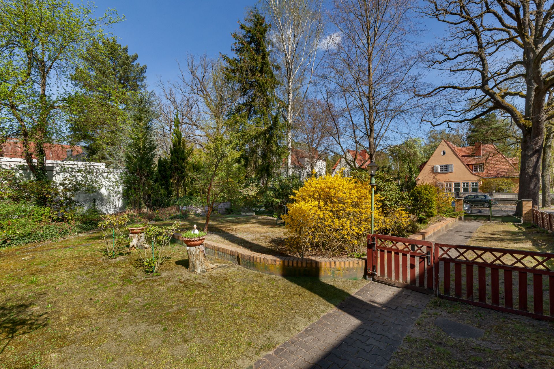 Garten mit Bäumen, Blumenbeeten, einem Zaun und einem kleinen Weg vor Wohnhäusern.