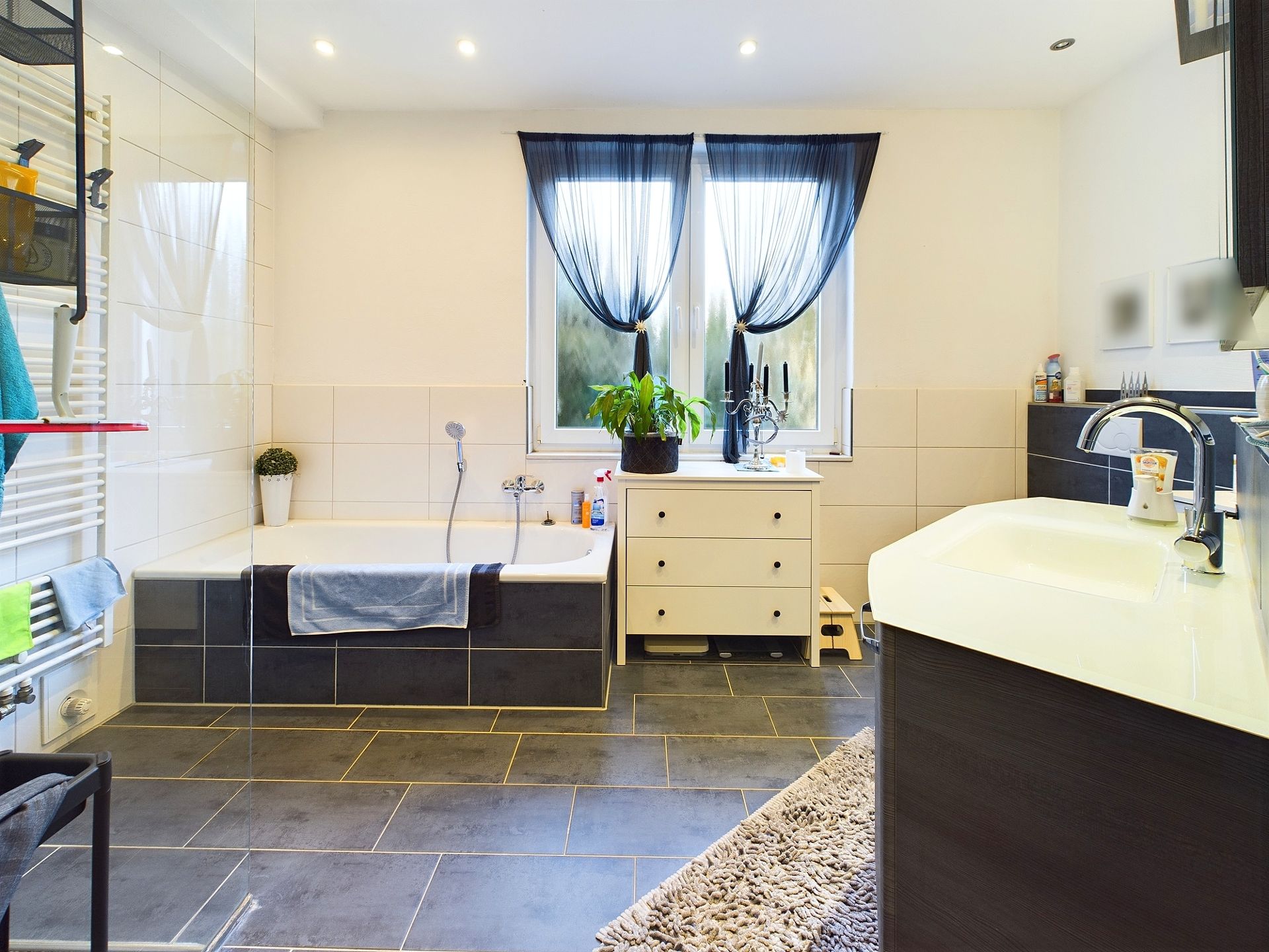 Modernes Badezimmer mit Badewanne, Waschbecken, Kommode, großen Fliesen und zwei Fenstern mit dunklen Vorhängen.