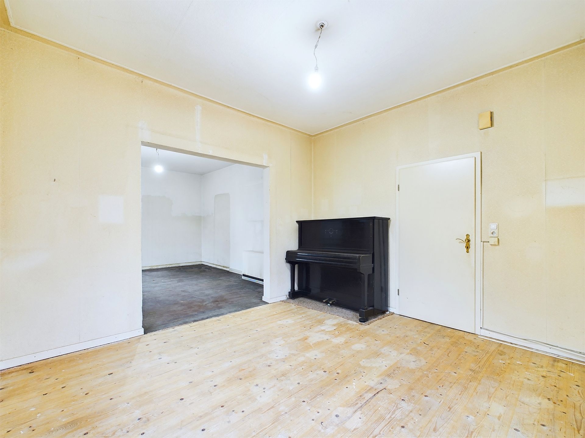 Helles Zimmer mit Holzboden, schwarzem Klavier und weißer Tür, angrenzender Raum sichtbar.