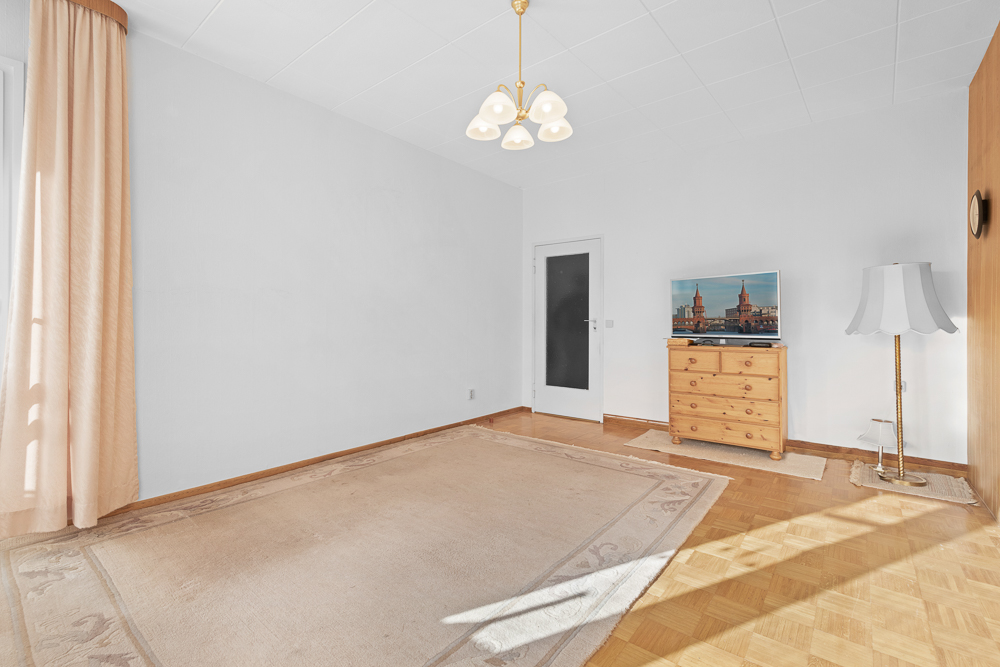 Helles Zimmer mit Teppich, Holzkommode, Stehlampe, TV und Vorhängen vor einem Fenster.