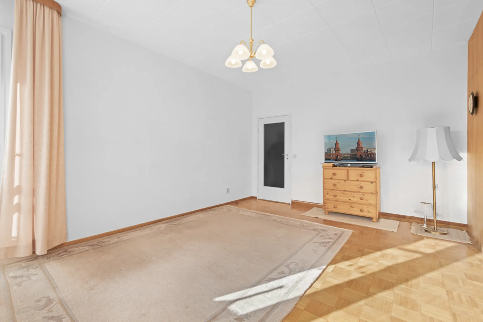 Helles Zimmer mit Teppich, Holzkommode, Stehlampe, TV und Vorhängen vor einem Fenster.