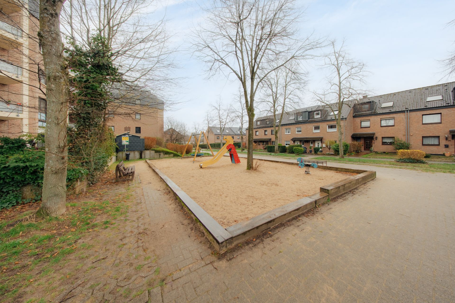 Spielplatz mit Sandfläche, Rutsche und Wippe zwischen Wohnhäusern, umgeben von Bäumen.