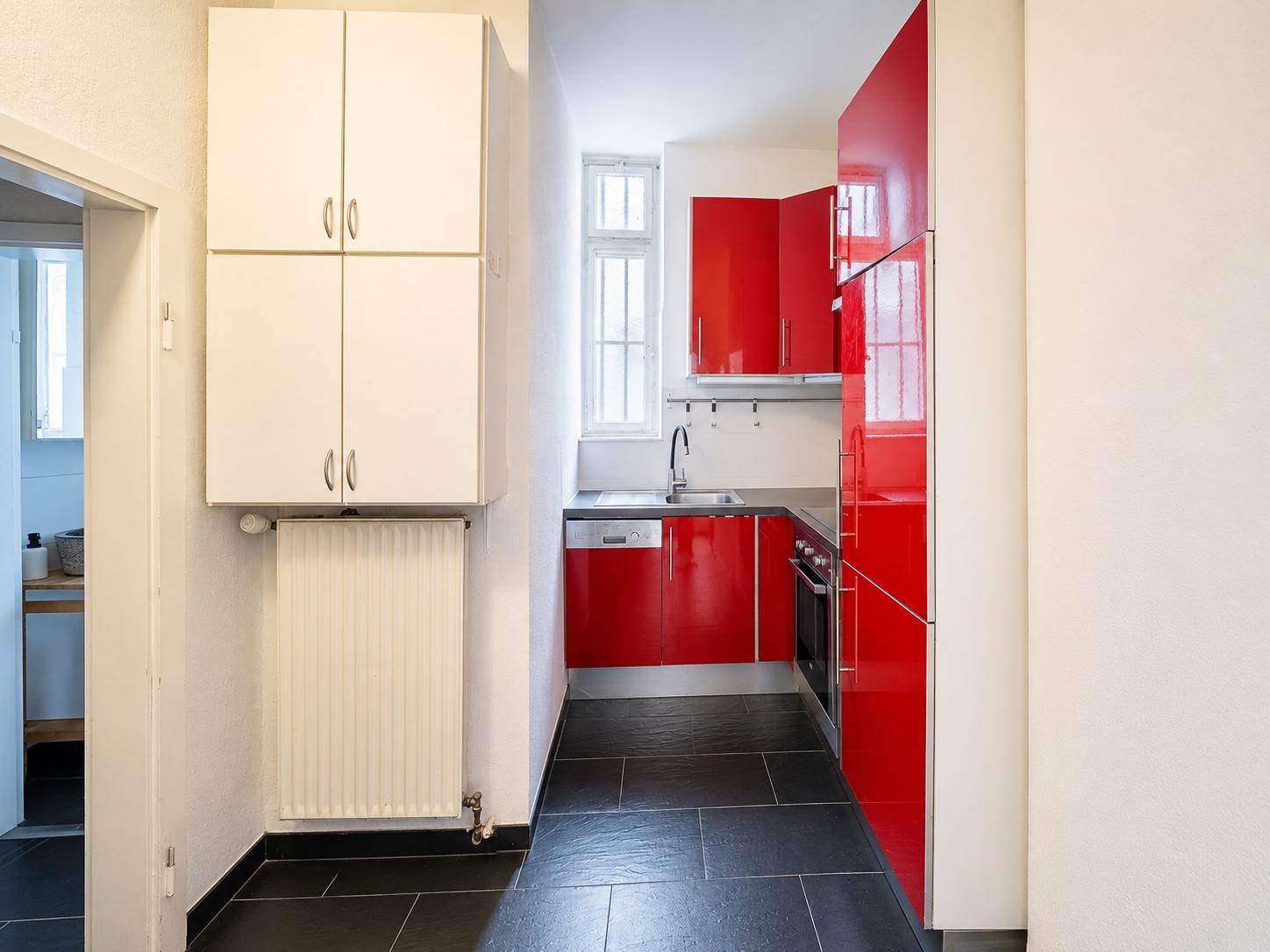 Schmale Küche mit roten Hochglanzschränken, weißem Hängeschrank und Fenster am Ende.