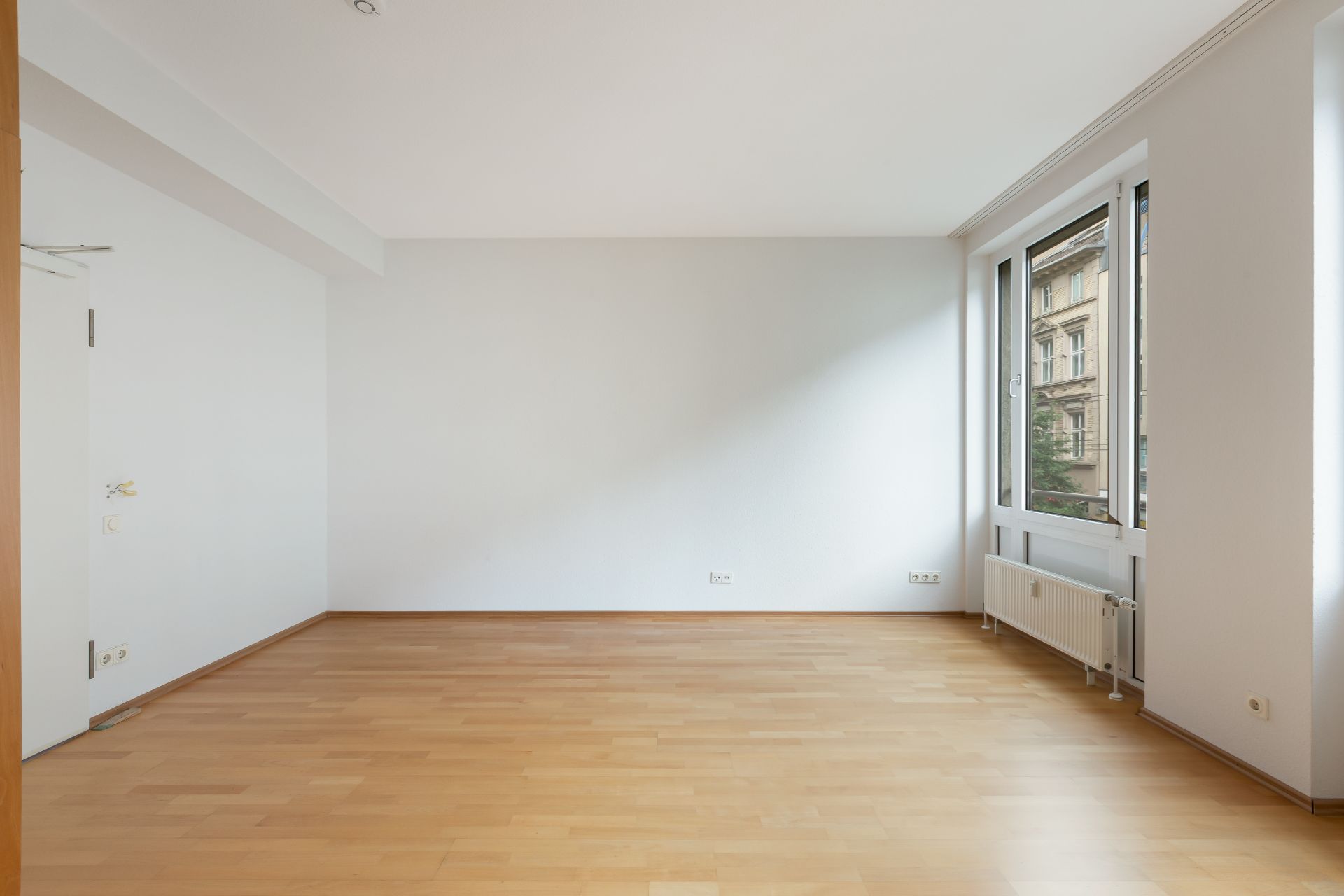 Leerer Raum mit hellem Holzboden, weißer Wand und großem Fenster mit Blick nach draußen.