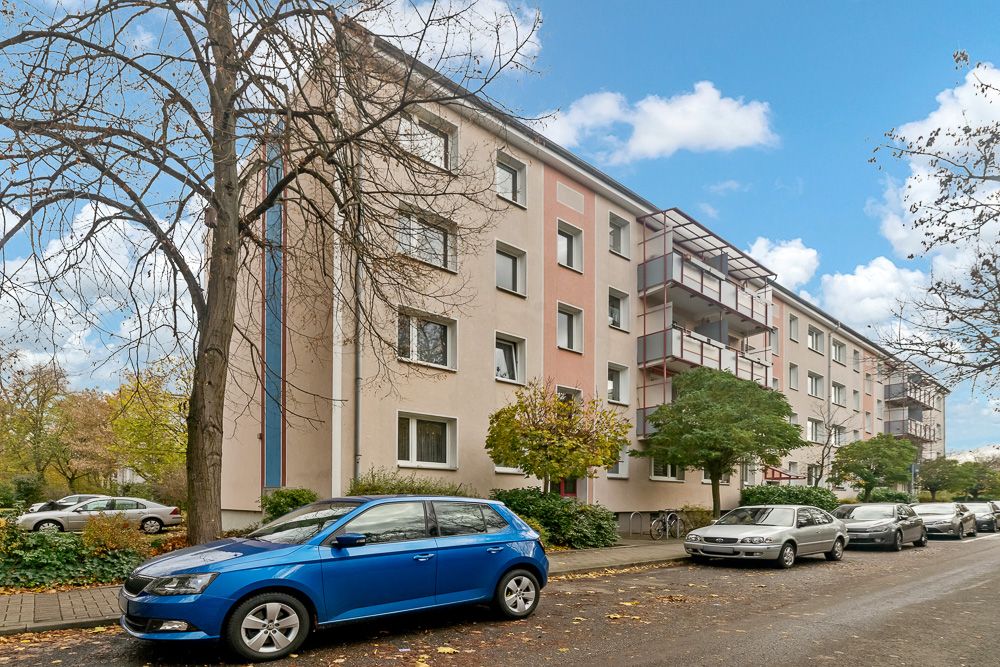 Mehrstöckiges Wohnhaus mit geparkten Autos und Bäumen am Straßenrand.
