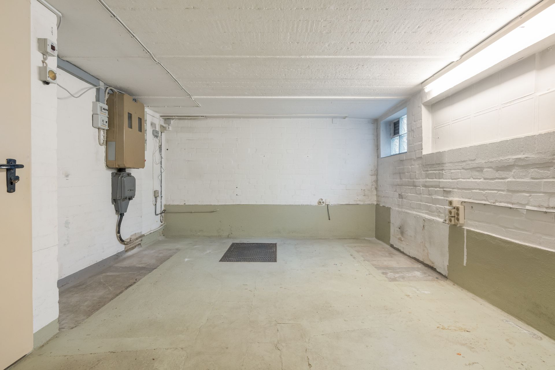 Leerer Kellerraum mit weißen Wänden, Fenster und sichtbaren Stromanschlüssen.