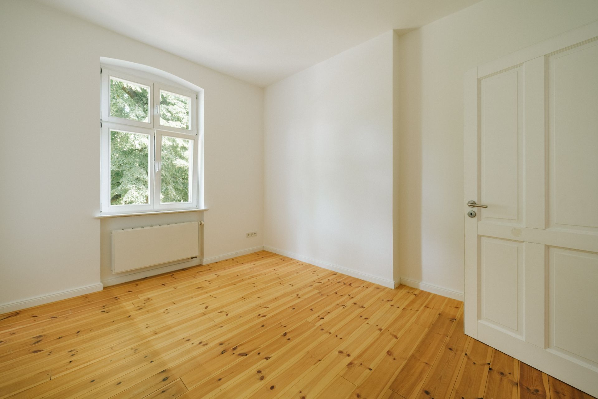 Leeres Zimmer mit Holzboden, weißer Tür und großem Fenster mit Blick auf Bäume.