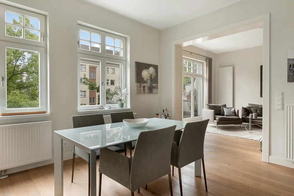 Esszimmer mit Glastisch und sechs Stühlen, dahinter Wohnzimmer mit Sofa, große Fenster.