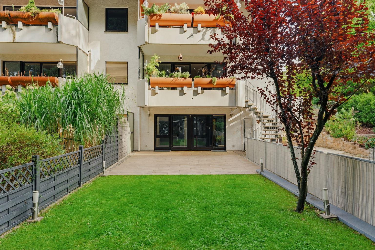 Garten mit Rasen, Terrasse und Baum vor einem Wohnhaus mit Balkonen.