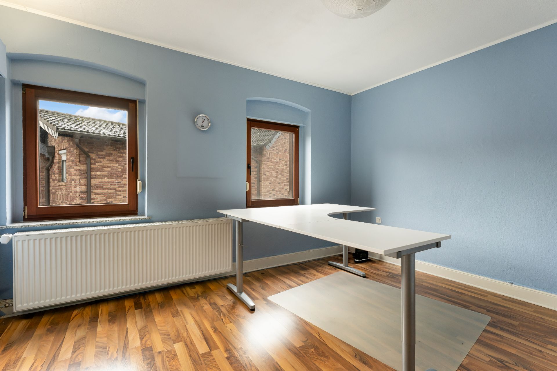Helles Zimmer mit zwei Fenstern, blau gestrichenen Wänden und einem weißen Schreibtisch auf Laminatboden.