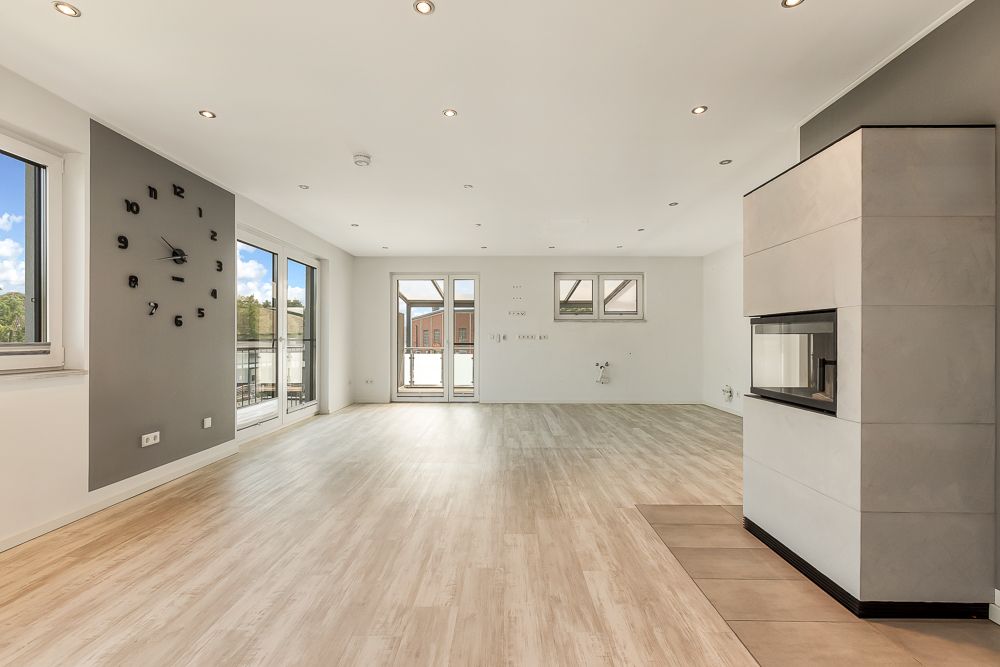 Heller, leerer Wohnraum mit Kamin, Holzfußboden, großen Fenstern und moderner Wanduhr.