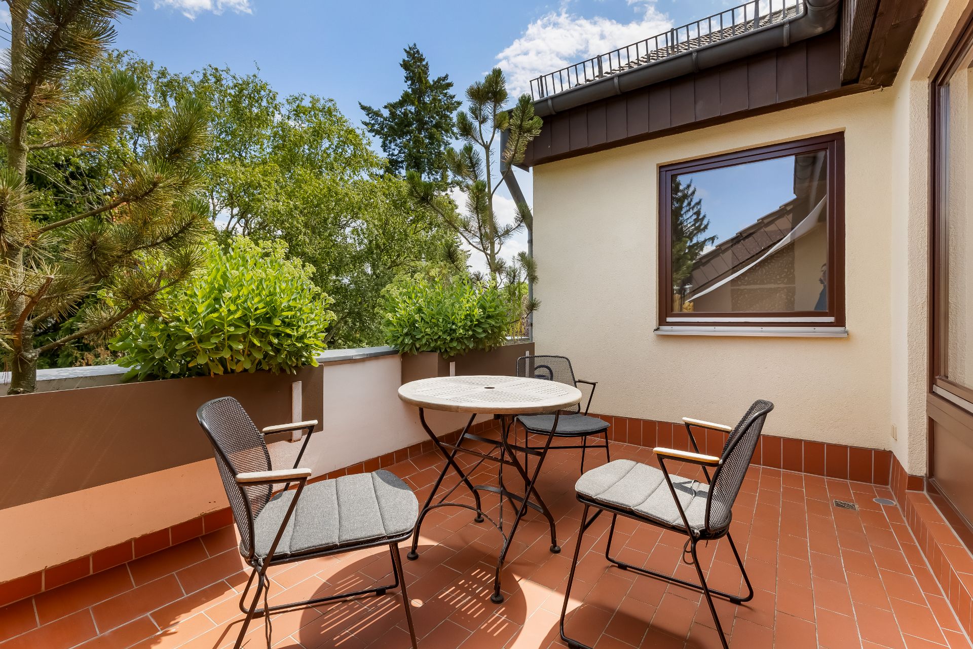 Terrasse mit rundem Tisch und vier Stühlen, umgeben von Pflanzen und Bäumen.