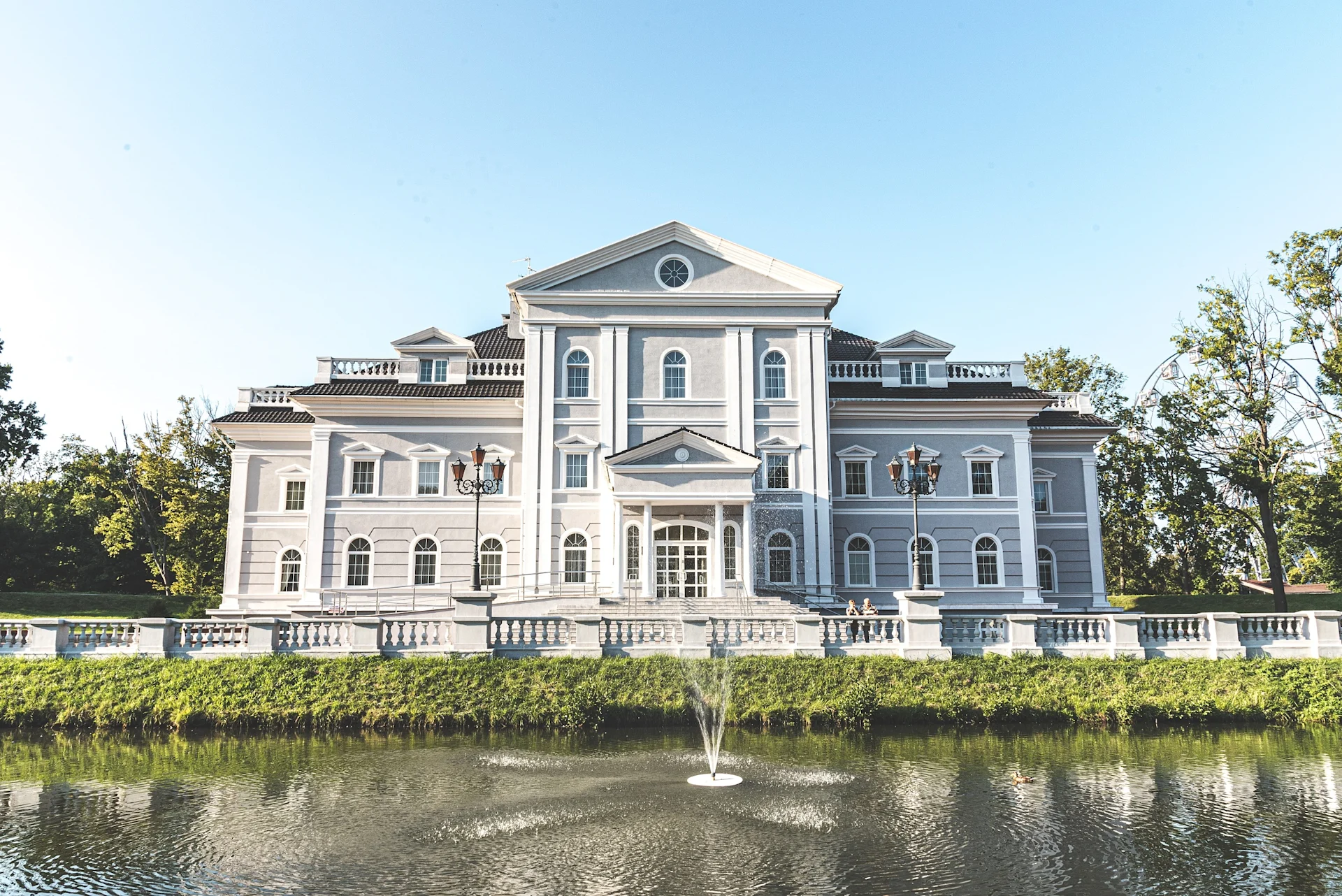 Großes, helles Wohngebäude im klassizistischen Stil mit Teich und Springbrunnen davor.