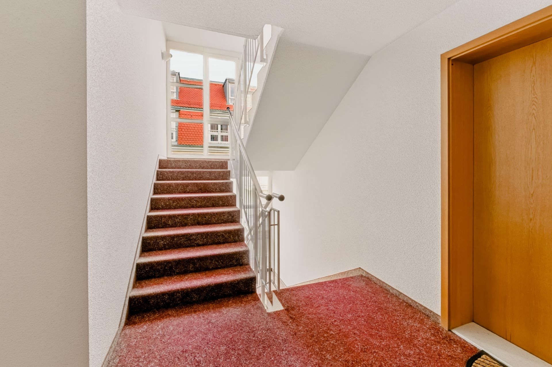 Treppenhaus mit rotem Teppichboden, Metallgeländer und Aufzugstür auf der rechten Seite.