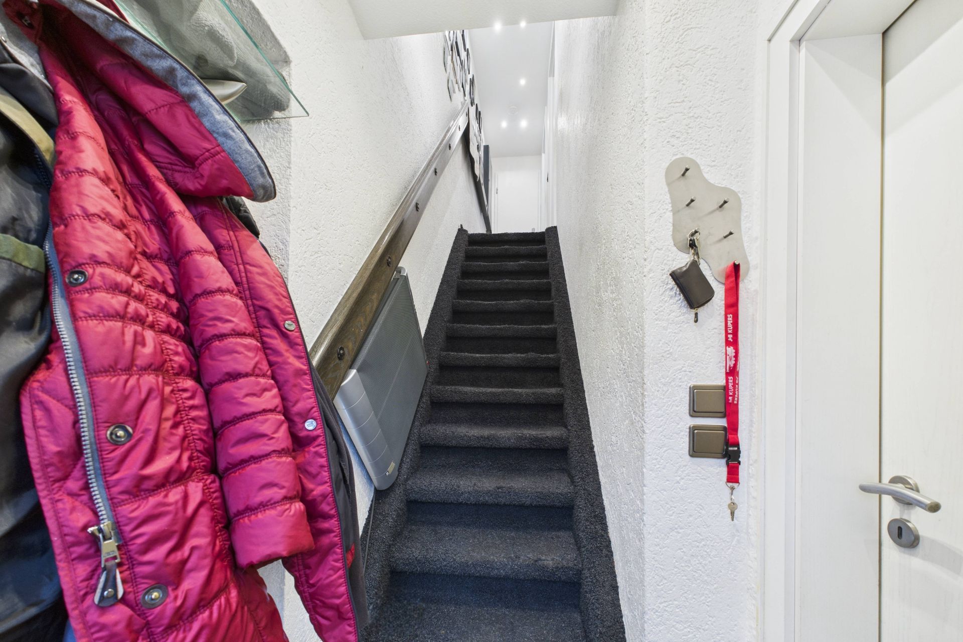 Treppenaufgang mit Teppich, links hängen Jacken, rechts Tür und Schlüsselbrett.