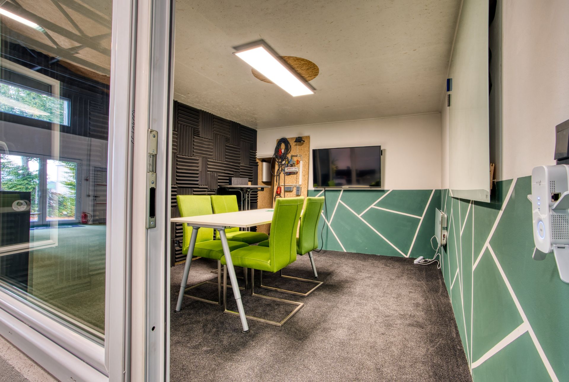 Kleiner Besprechungsraum mit grünem Stuhlset, Wandtafel und Bildschirm an der Wand.