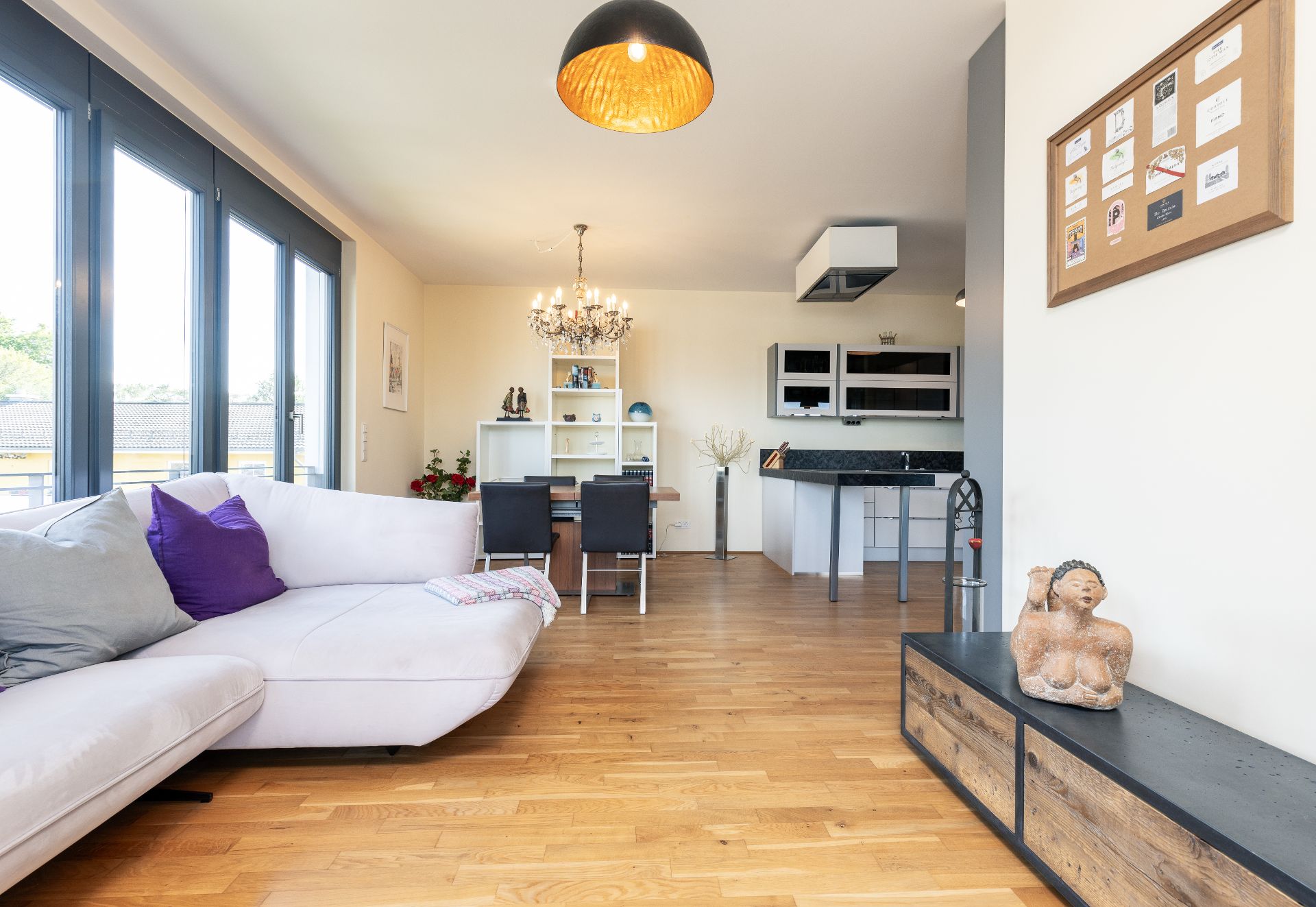 Helles Wohnzimmer mit Ecksofa, Holzboden, Esstisch und offener Küche.