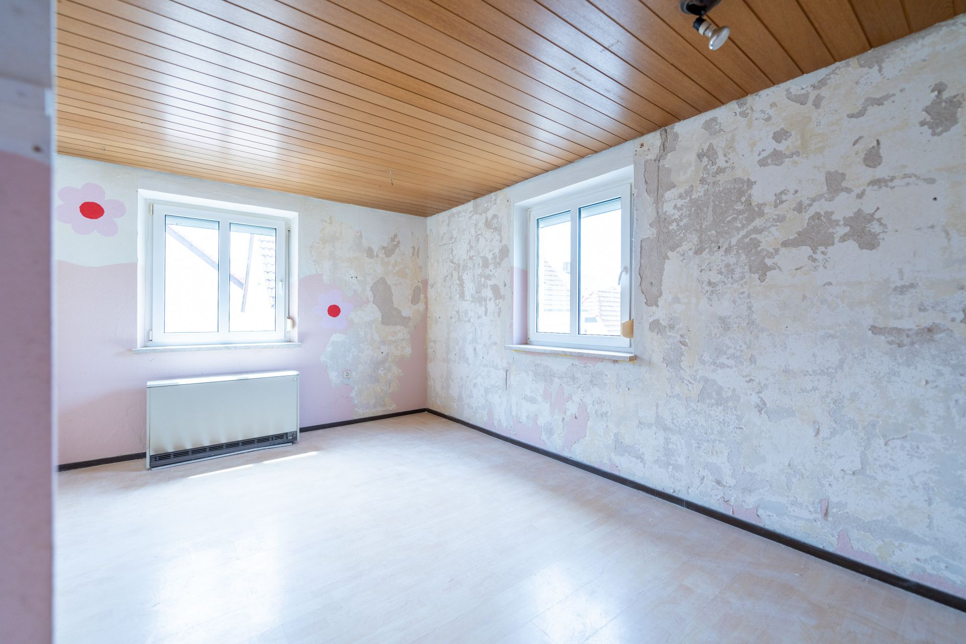 Leeres Zimmer mit zwei Fenstern, Holzdecke und renovierungsbedürftigen Wänden.
