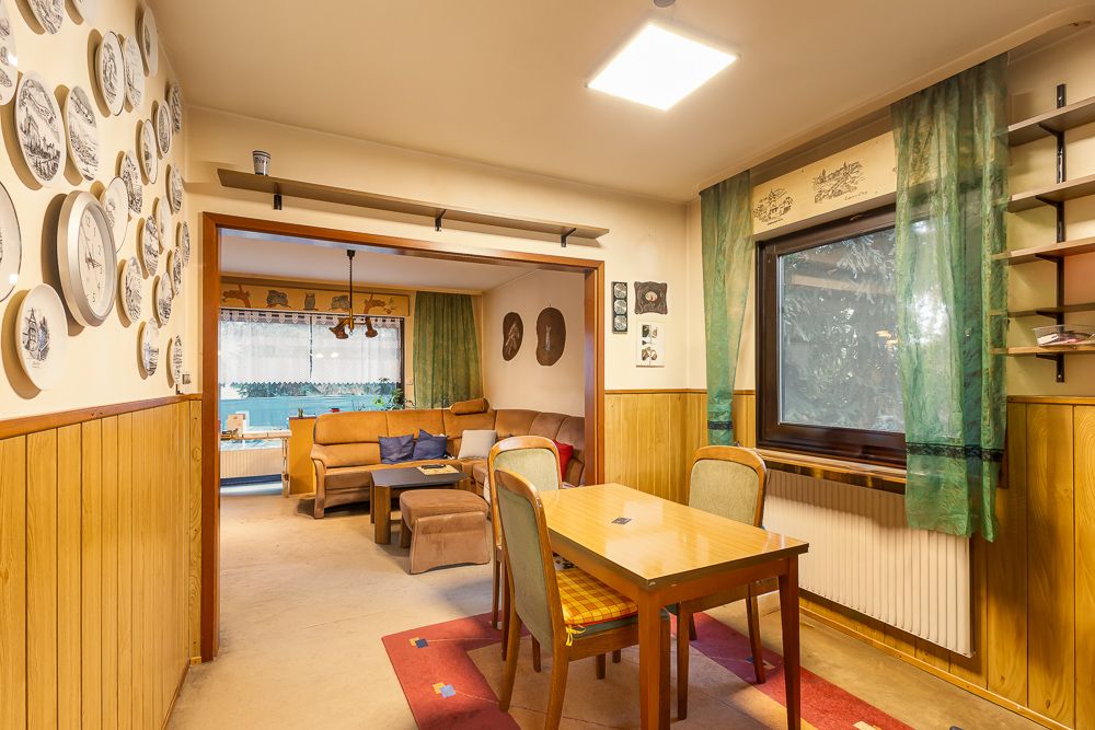 Esszimmer mit Holztisch, Stühlen, grünen Vorhängen und Wandtellern, dahinter ein Wohnzimmer mit Sofa.