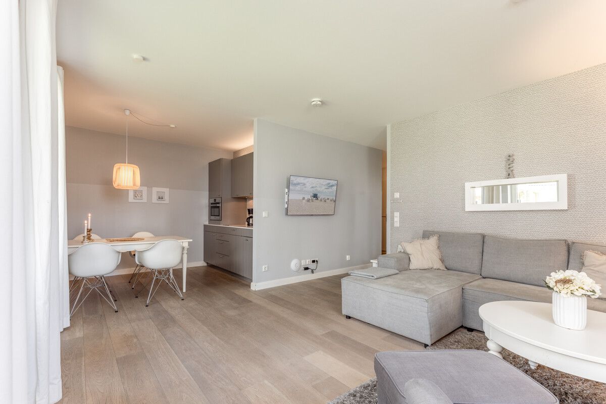 Helles Wohnzimmer mit grauem Sofa, rundem Tisch und offener Küche mit Essbereich.