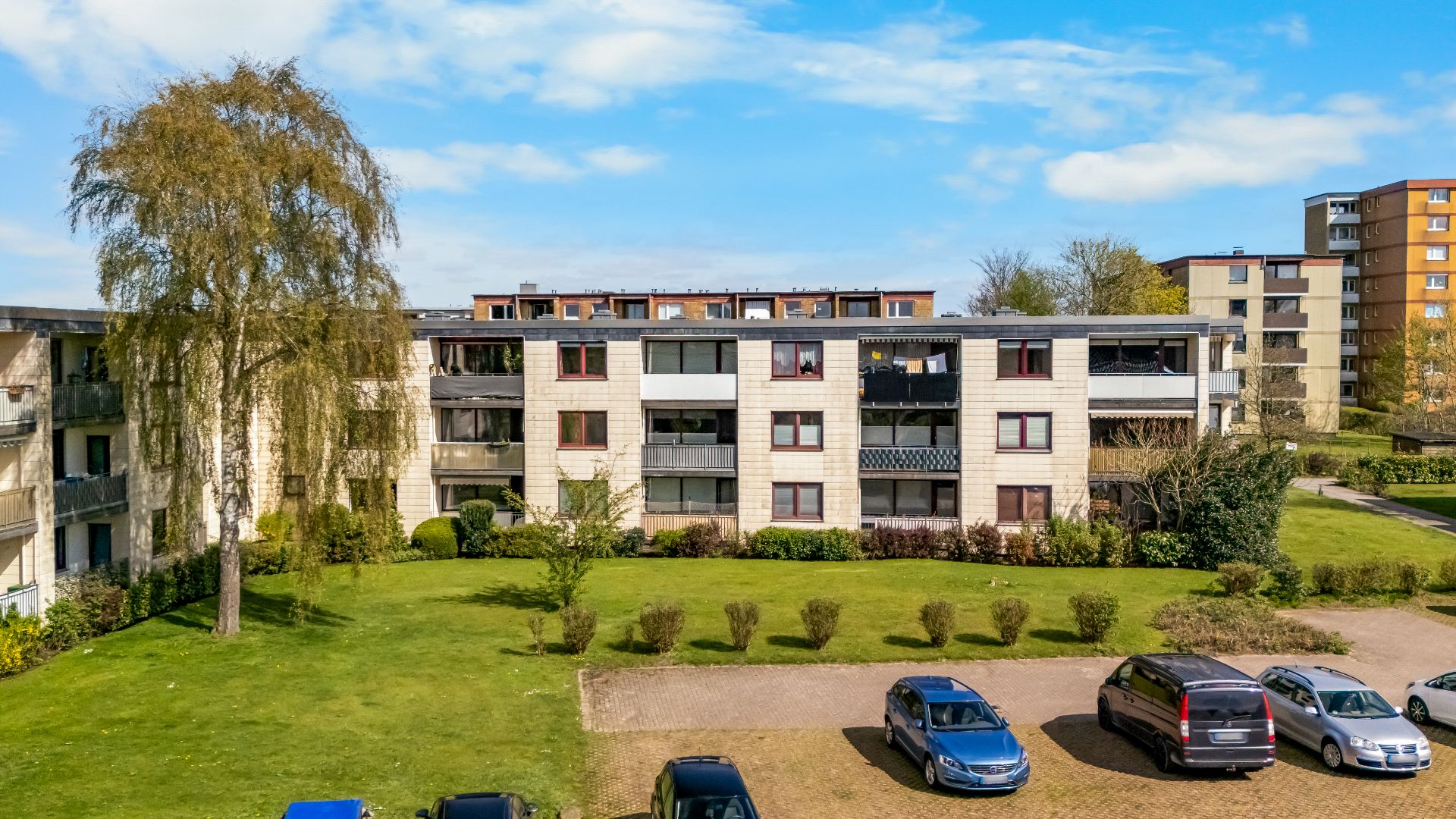 Mehrfamilienhaus mit Balkonen, davor eine Rasenfläche und parkende Autos auf einem Parkplatz.