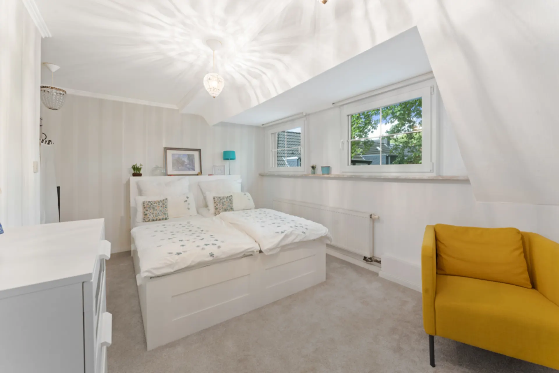 Helles Schlafzimmer mit Doppelbett, gelbem Sessel und zwei Fenstern.