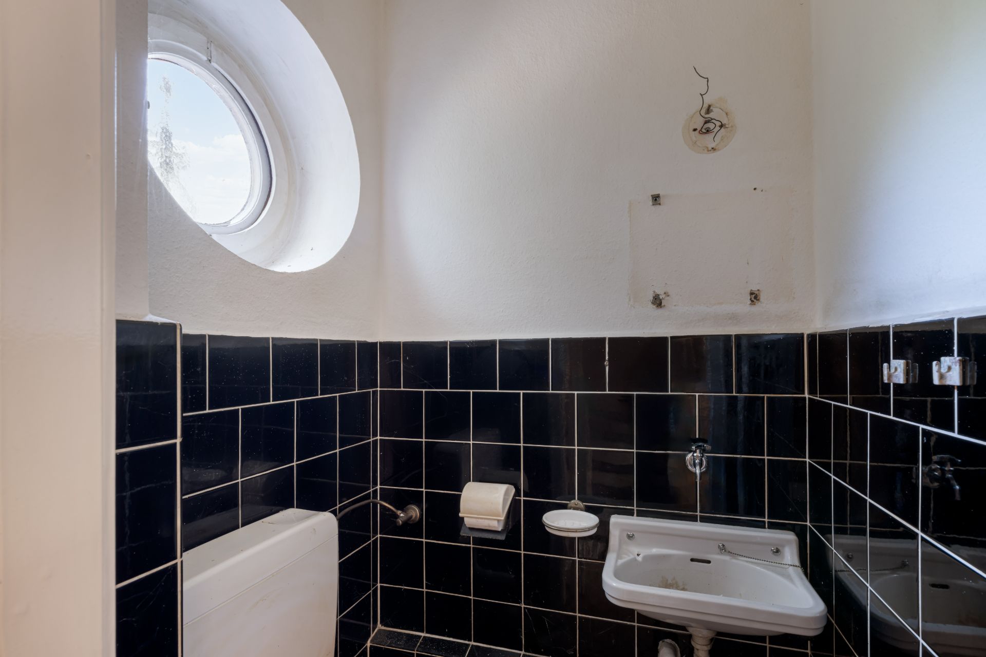 Kleines Bad mit rundem Fenster, schwarzer Fliesenwand, Waschbecken und Toilettenpapierhalter.