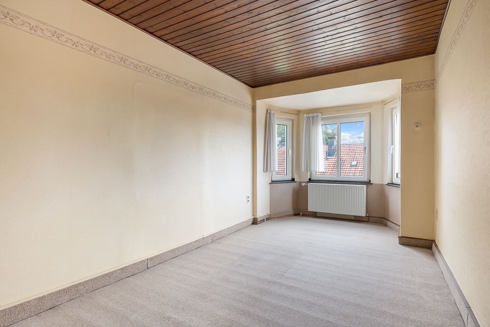 Leeres Zimmer mit Teppichboden, Holzdecke und Erkerfenster mit Heizung darunter.