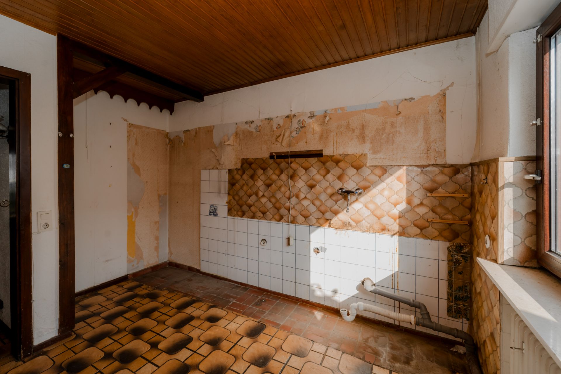 Leeres Zimmer mit alten Wandfliesen, Rohrleitungen und braunem Fliesenboden.