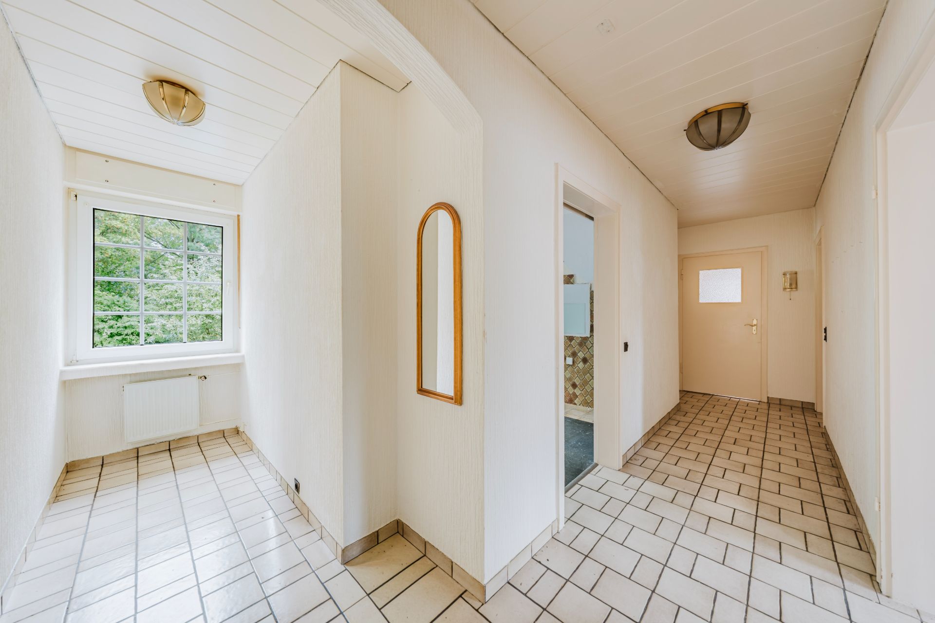 Heller Flur mit Fliesenboden, Fenster am Ende, Türen zu mehreren Zimmern und einem Wandspiegel.