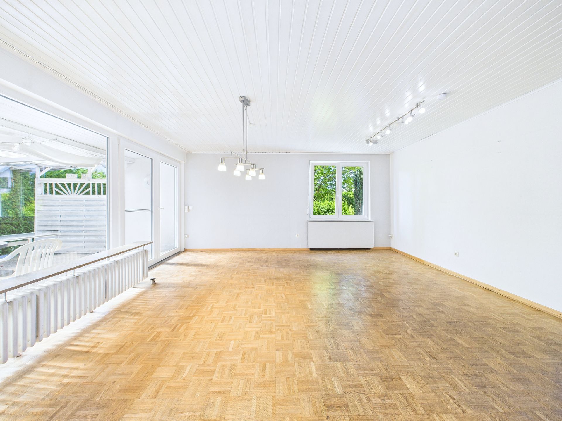 Heller, leerer Raum mit Parkettboden, weißen Wänden, Deckenleuchten und großen Fenstern.