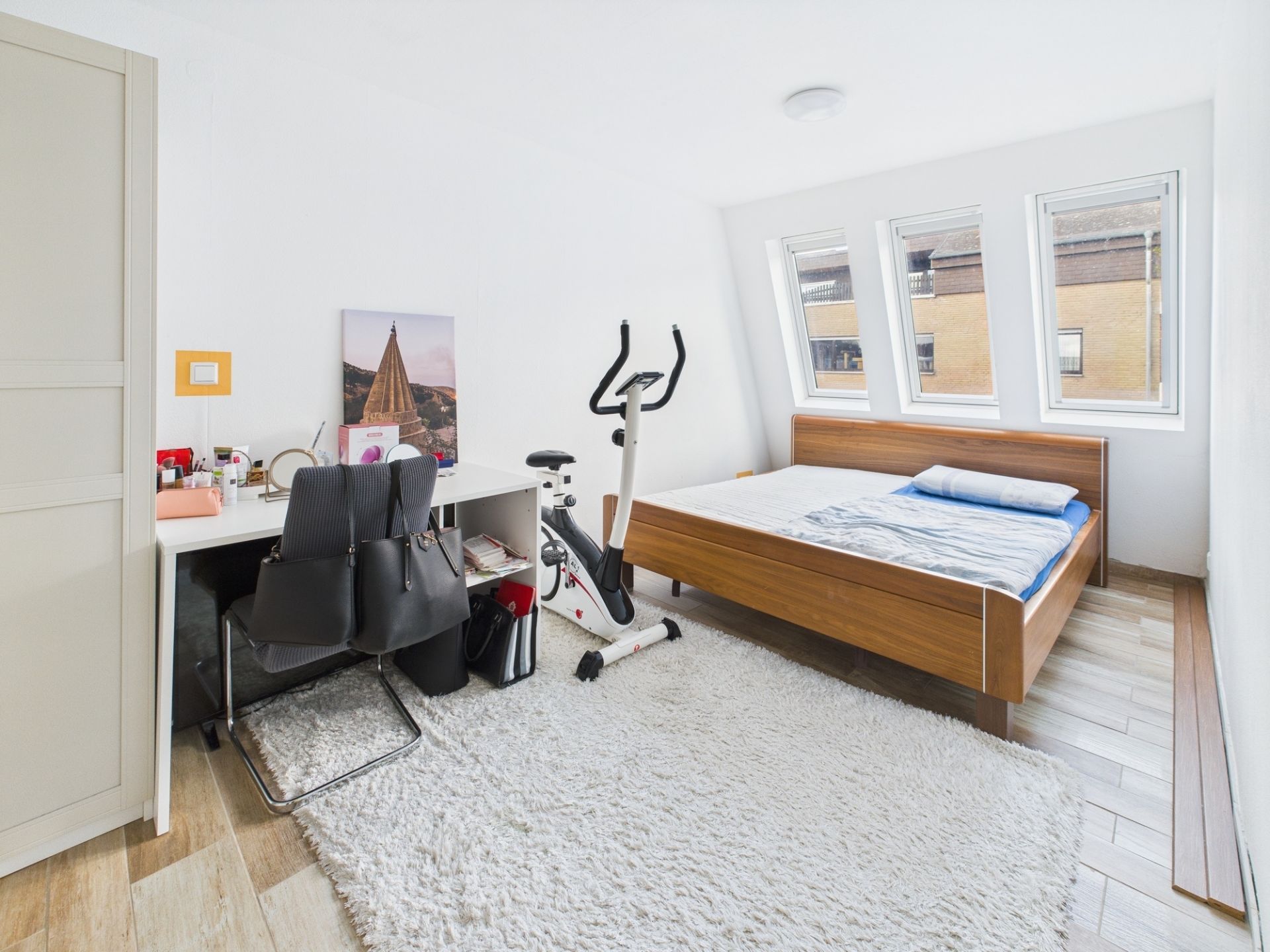 Helles Schlafzimmer mit Schreibtisch, Stuhl, Heimtrainer, Bett und drei Fenstern.