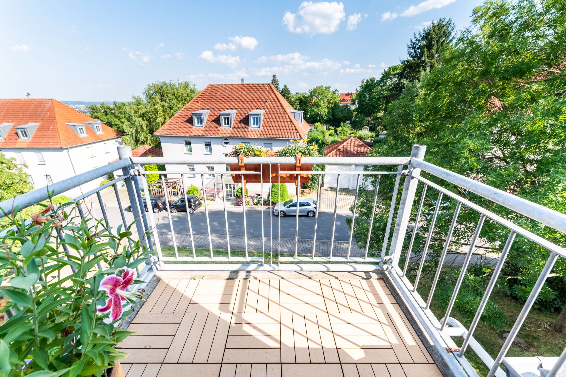 Balkon mit Holzboden und Pflanzen, Blick auf Häuser mit roten Dächern und Bäume.