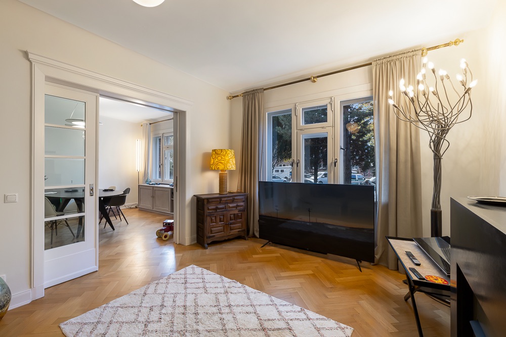 Helles Wohnzimmer mit Teppich, TV, Lampe, Holzboden und großen Fenstern, angrenzendes Esszimmer sichtbar.