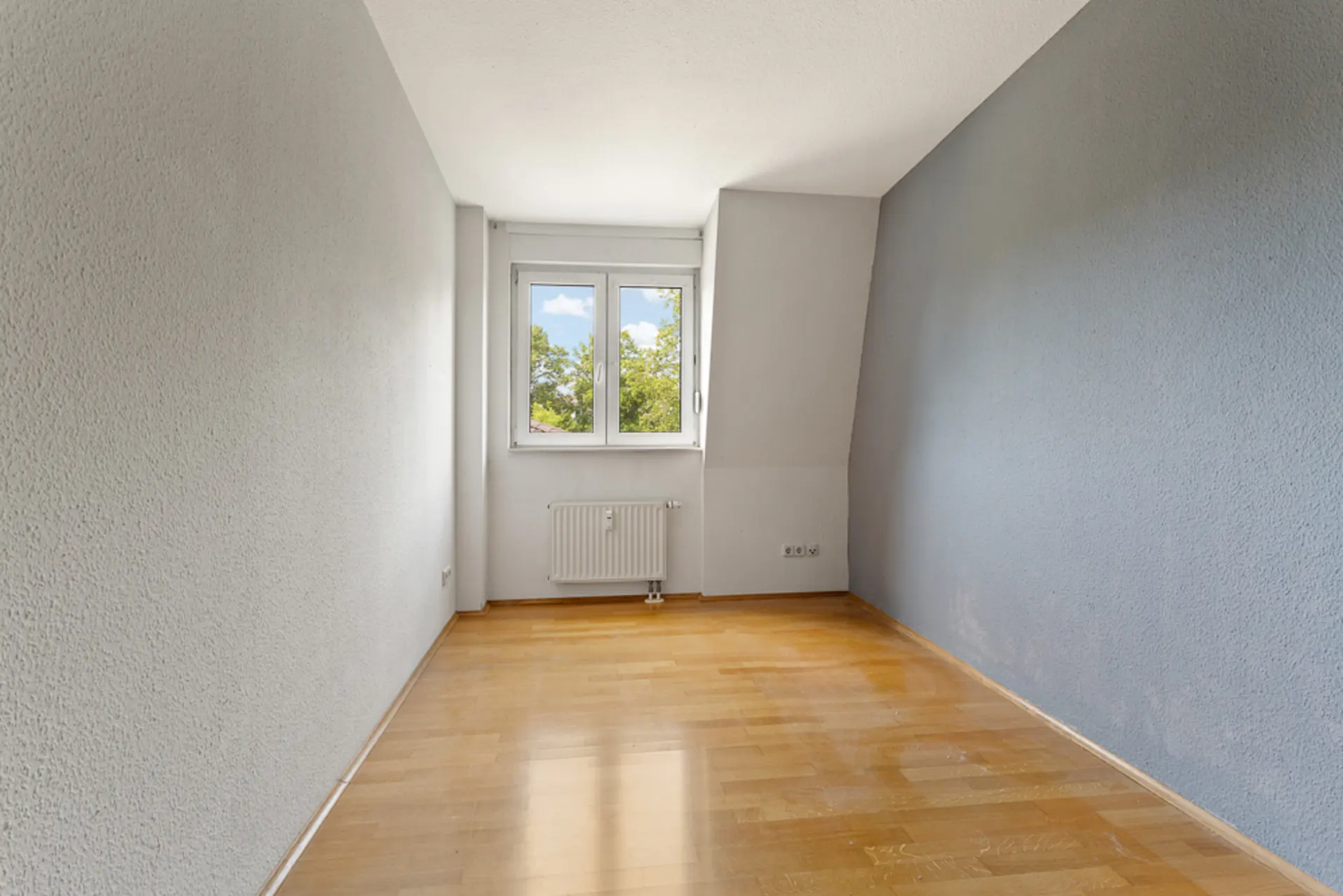 Leerer Raum mit Holzboden, grauen Wänden und Fenster mit Blick ins Grüne.