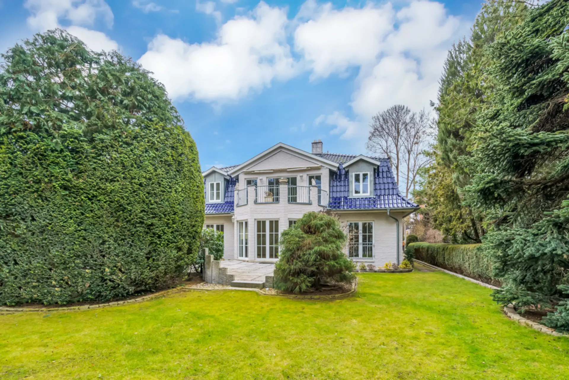 Zweistöckiges Wohnhaus mit blauem Dach, großem Garten und Bäumen an den Seiten.