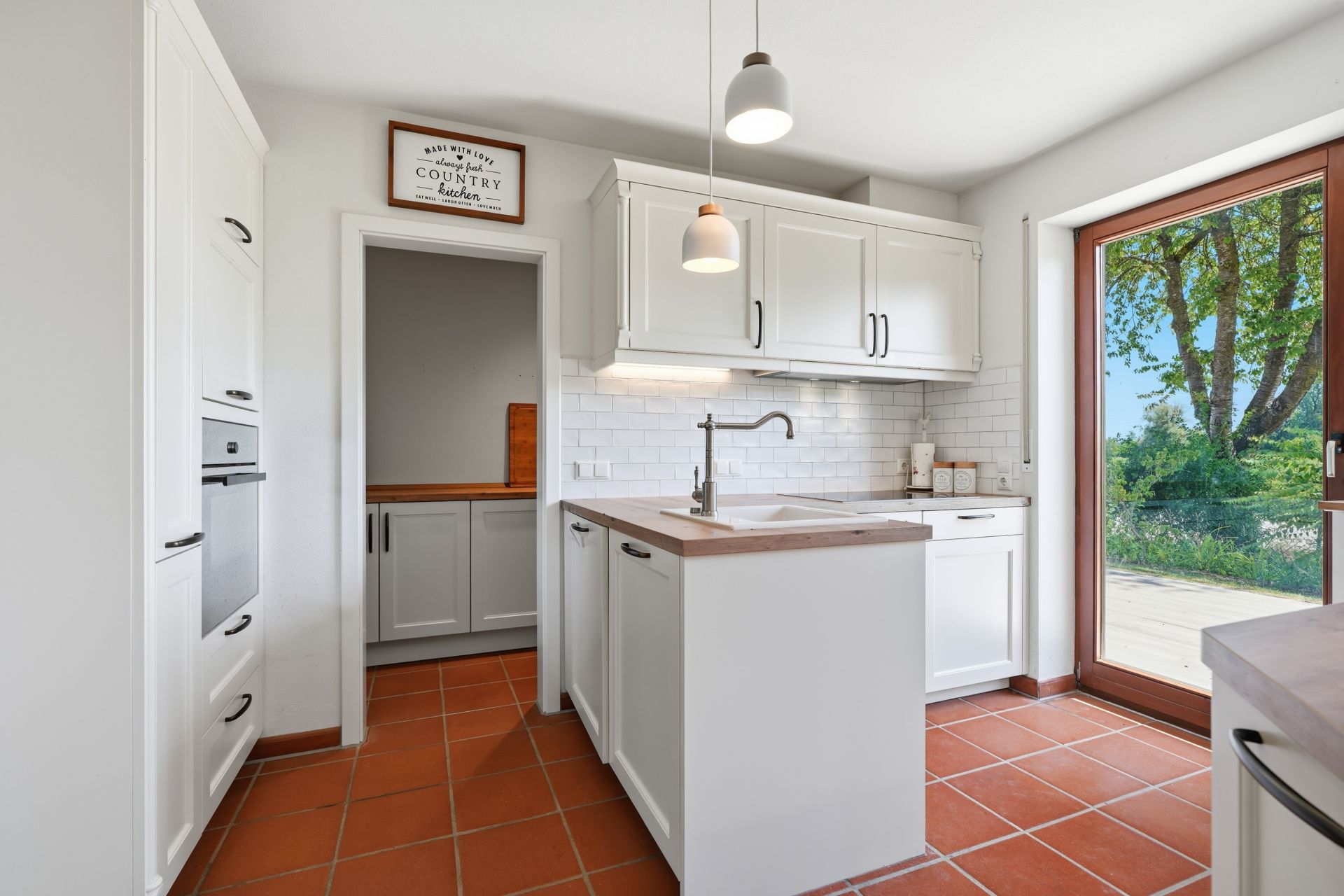Helle Küche mit weißen Schränken, Kücheninsel, Fliesenboden und großem Fenster zur Terrasse.